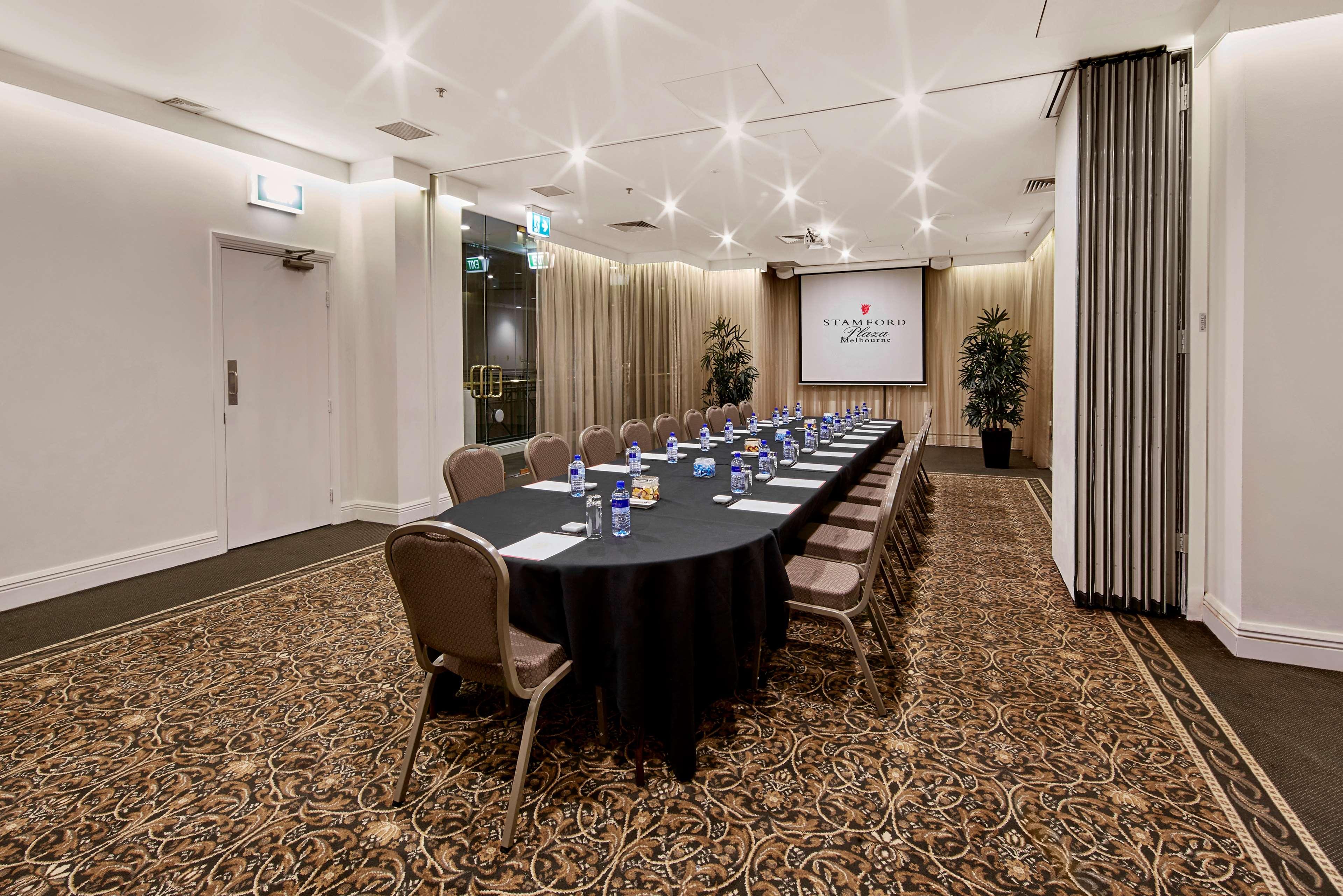 Stamford Plaza Melbourne Hotell Exteriör bild The photo shows a meeting room set up for a conference or presentation. There is a long, rectangular table covered with a black tablecloth, surrounded by chairs. Each seat has a water bottle placed in front of it. At the front of the room, there is a