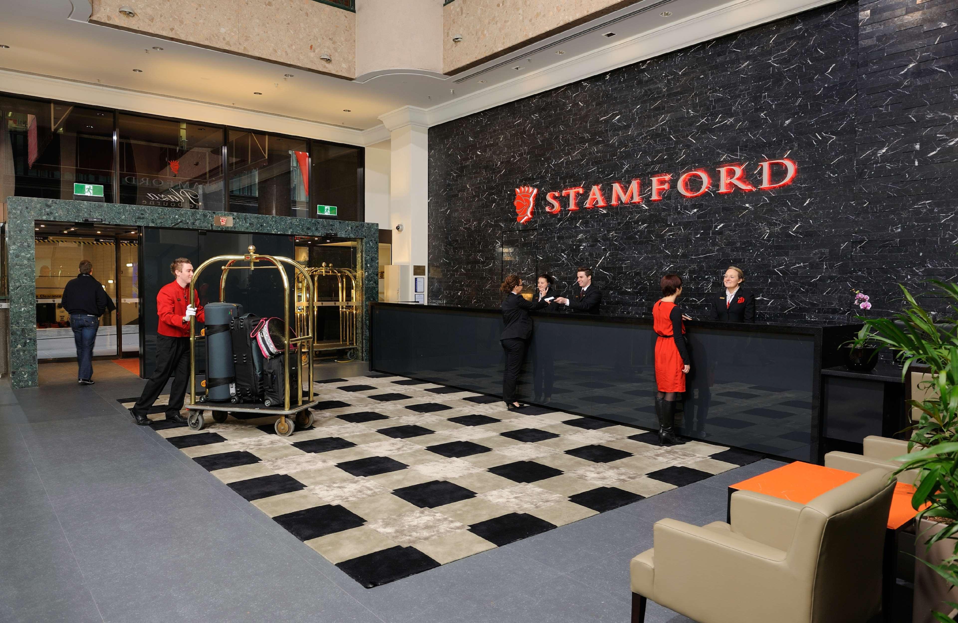 Stamford Plaza Melbourne Hotell Inreriör bild The photo shows the interior of a hotel lobby, specifically the Stamford Hotel. There is a large reception desk where several staff members are attending to guests. In the foreground, a bell cart loaded with luggage is positioned beside the reception