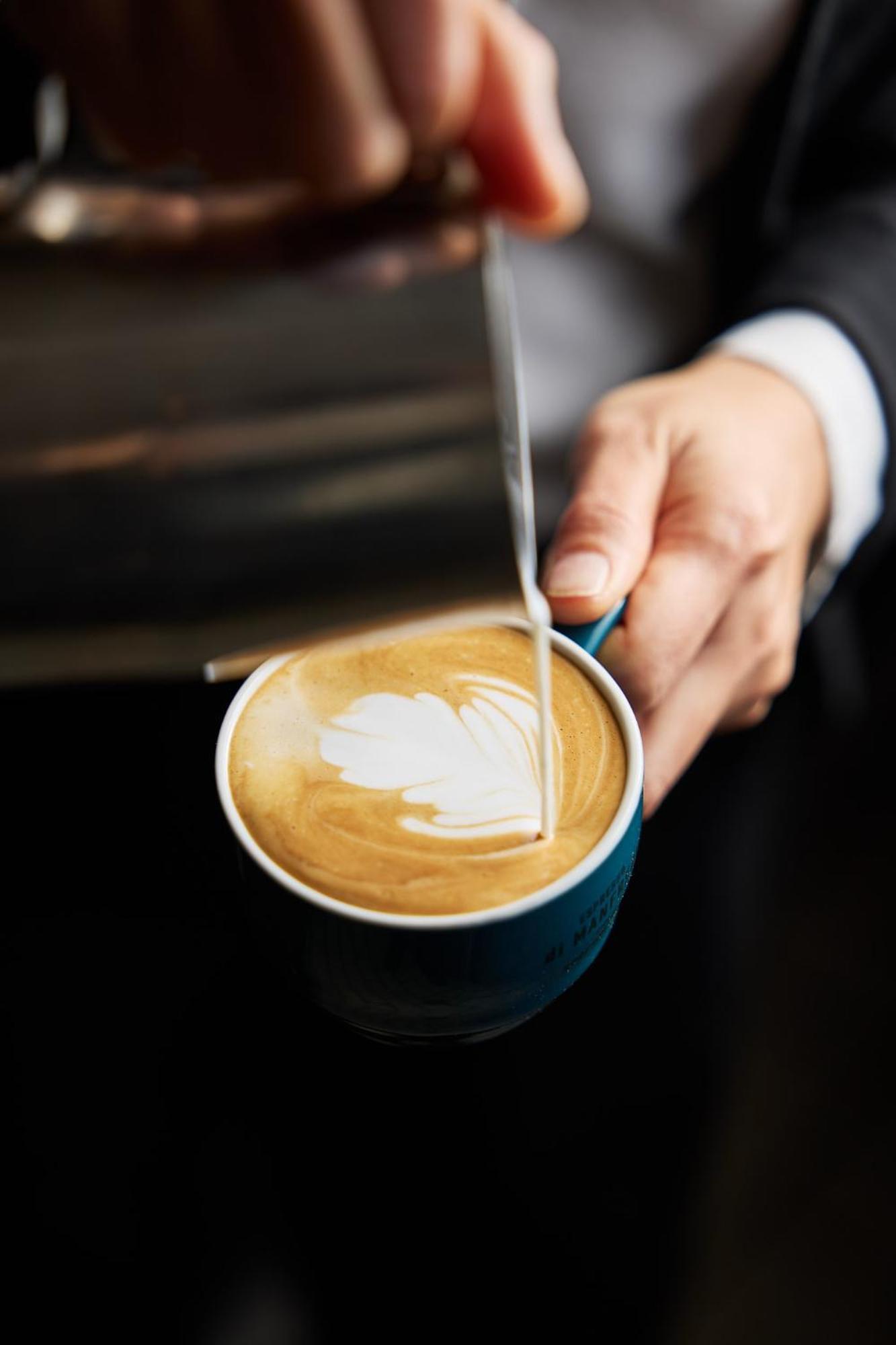 Stamford Plaza Melbourne Hotell Exteriör bild The photo shows a close-up of a hand holding a cup of coffee with a latte art design on the surface. The coffee appears to be a creamy, caramel color, likely a latte or cappuccino. A small stream of milk is being poured into the cup, creating a heart