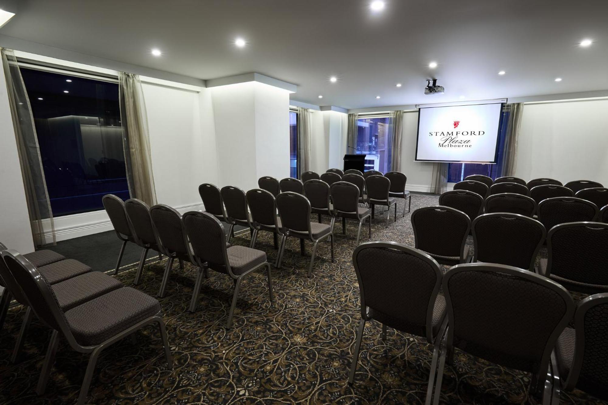 Stamford Plaza Melbourne Hotell Exteriör bild The image shows a conference or meeting room set up for an event. There are rows of gray chairs arranged facing a large screen displaying the name "Warford House." The room is well-lit with overhead lighting and has large windows covered with light-c