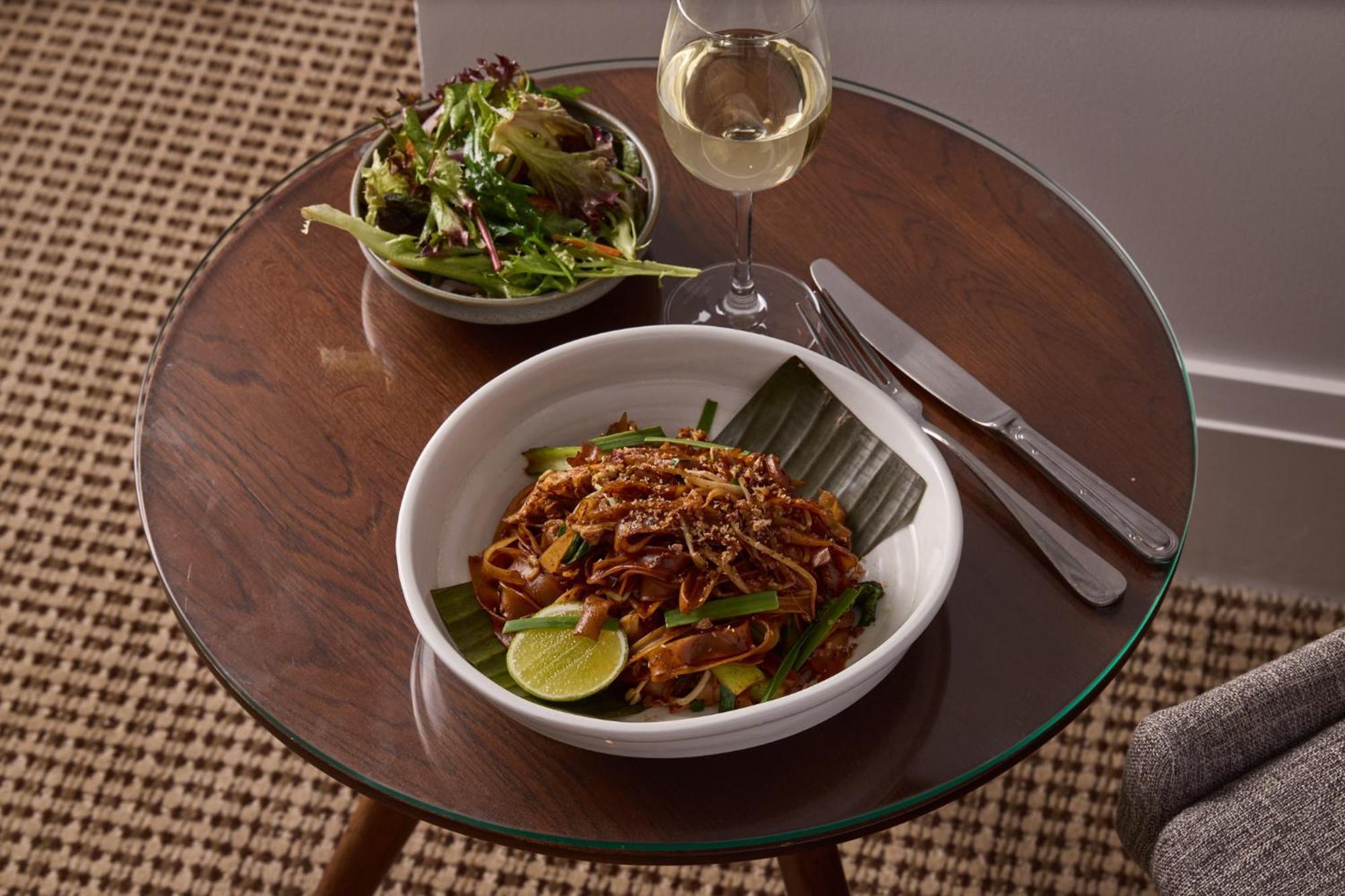 Stamford Plaza Melbourne Hotell Exteriör bild The photo shows a meal served on a wooden table. In the foreground, there is a bowl of stir-fried noodles garnished with green onions and a lime wedge, set atop a banana leaf. Next to it, there is a bowl of mixed green salad. A glass of white wine si