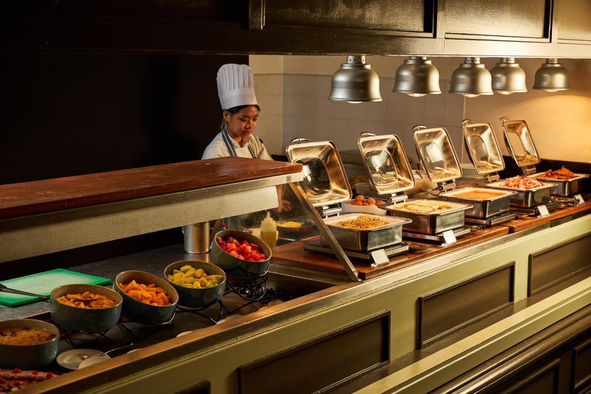 Stamford Plaza Melbourne Hotell Exteriör bild The photo shows a buffet setup in a dining area. There is a chef in a white uniform and hat, standing behind a counter filled with various food options. The buffet features several metal serving trays containing different dishes, with warming lights 